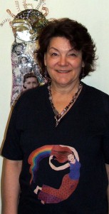 Photo of Tammy Vitale is wearing Tangerine Meg Rainbow Lady tshirt design, along with one of her own necklaces, in front of her own beautiful sculpture