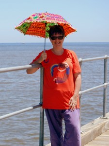 Yvette J on the jetty, she is smiling and wearing Rainbow Lady tshirt design on orange shirt, and holding a rainbow umbrella.