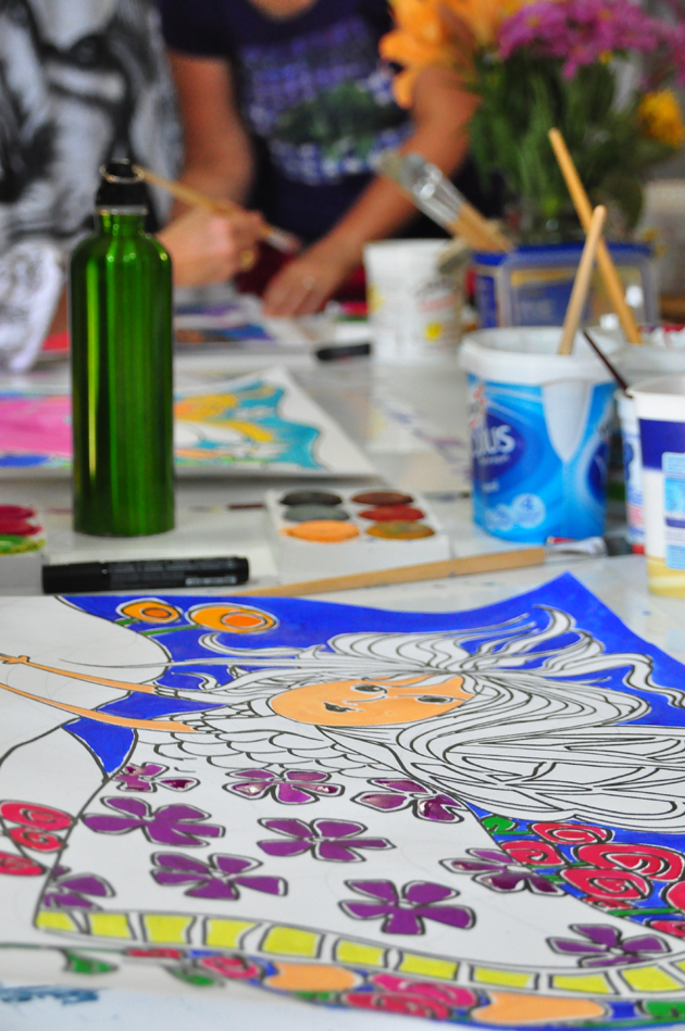 photographic view along the happily cluttered table at art workshop, showing water pots, drinking water, students work and paints, looking along to the leader