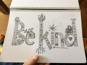 left hand holding a page of a spiral bound book with "Be Kind" typographic illustration marker drawing