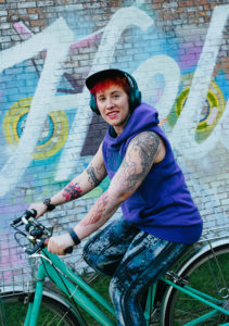 MIchelle Nickolaisen riding a green bike in front of a graffiti wall that says "Hello"